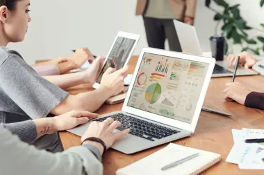 Osoby pracują przy stole z laptopami i tabletem; na ekranie jednego laptopa widać wykresy.