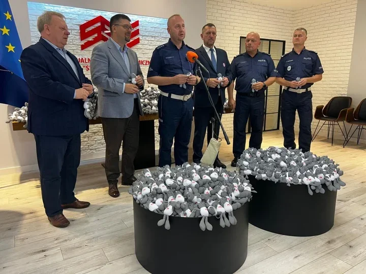 Grupa mężczyzn, w tym policjanci i urzędnicy, stoi podczas konferencji prasowej obok pojemników wypełnionych pluszowymi słonikami.