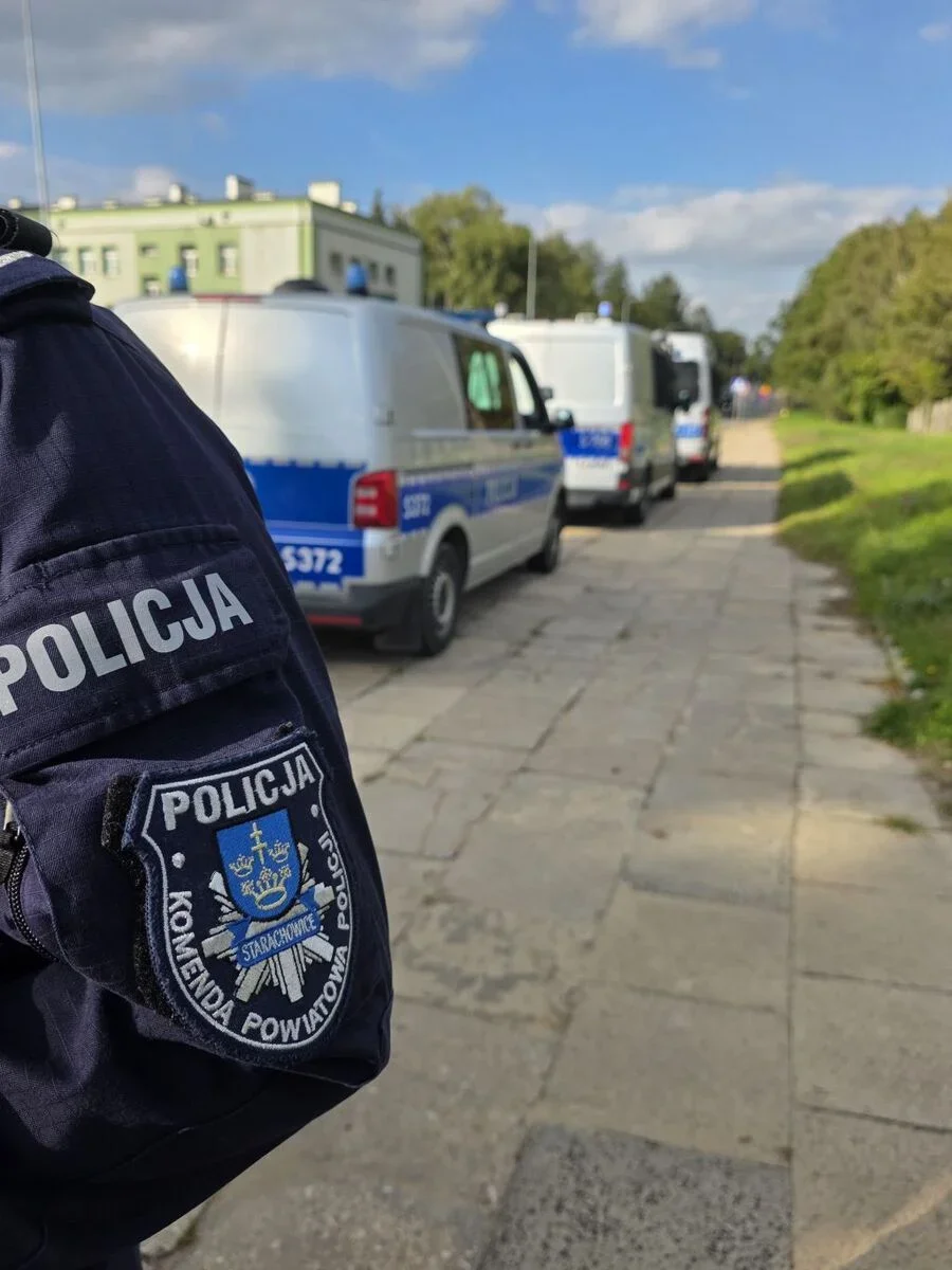 Funkcjonariusz policji z komendy powiatowej w Starachowicach stoi przy zaparkowanych radiowozach na brukowanej drodze w słoneczny dzień.