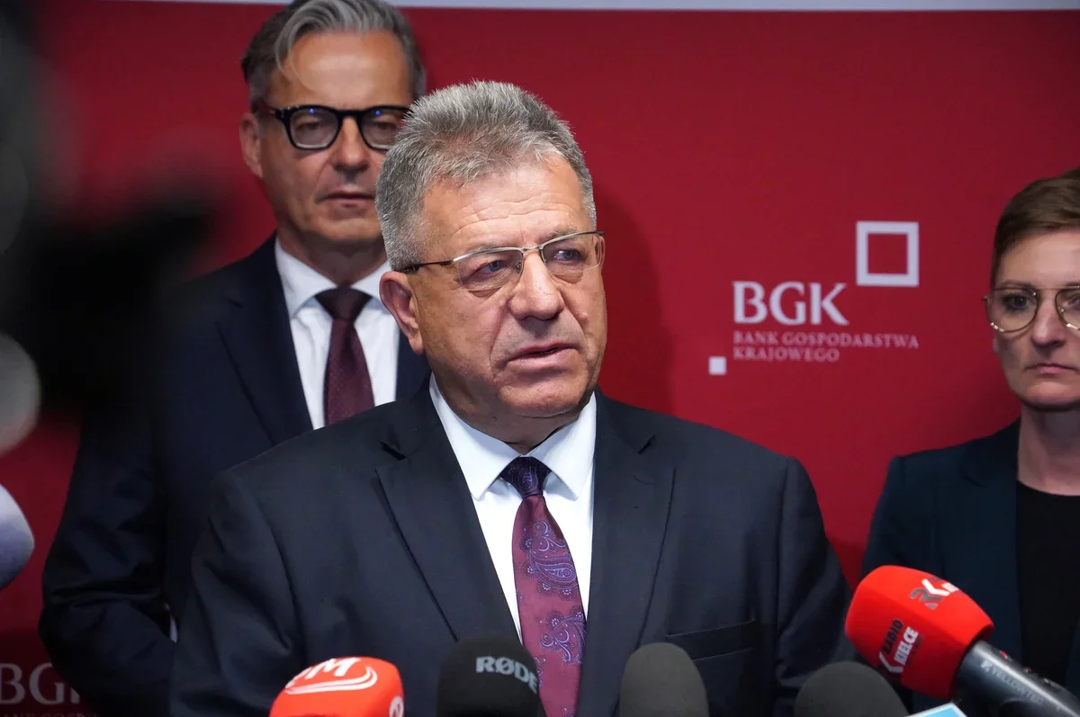 Mężczyzna w garniturze przemawia podczas konferencji prasowej przed czerwonym tłem z logo BGK, otoczony mikrofonami mediów.