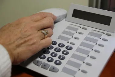 Dłoń osoby z pierścionkiem naciska klawisze białego telefonu biurowego leżącego na drewnianym blacie.