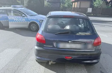 Policyjny radiowóz z napisem POLICJA zatrzymał ciemny samochód osobowy marki Peugeot na drodze w zabudowie jednorodzinnej.