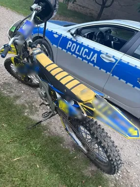 Motocykl crossowy stoi obok policyjnego radiowozu oznaczonego napisem POLICJA na szutrowej drodze.
