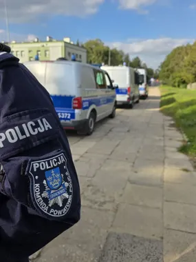Funkcjonariusz policji z komendy powiatowej w Starachowicach stoi przy zaparkowanych radiowozach na brukowanej drodze w słoneczny dzień.