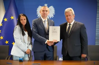 Trzy osoby w gabinecie z flagą UE i godłem na niebieskim tle, dwaj mężczyźni podają sobie ręce i trzymają dokument z napisem „Wojewoda Świętokrzyski”.
